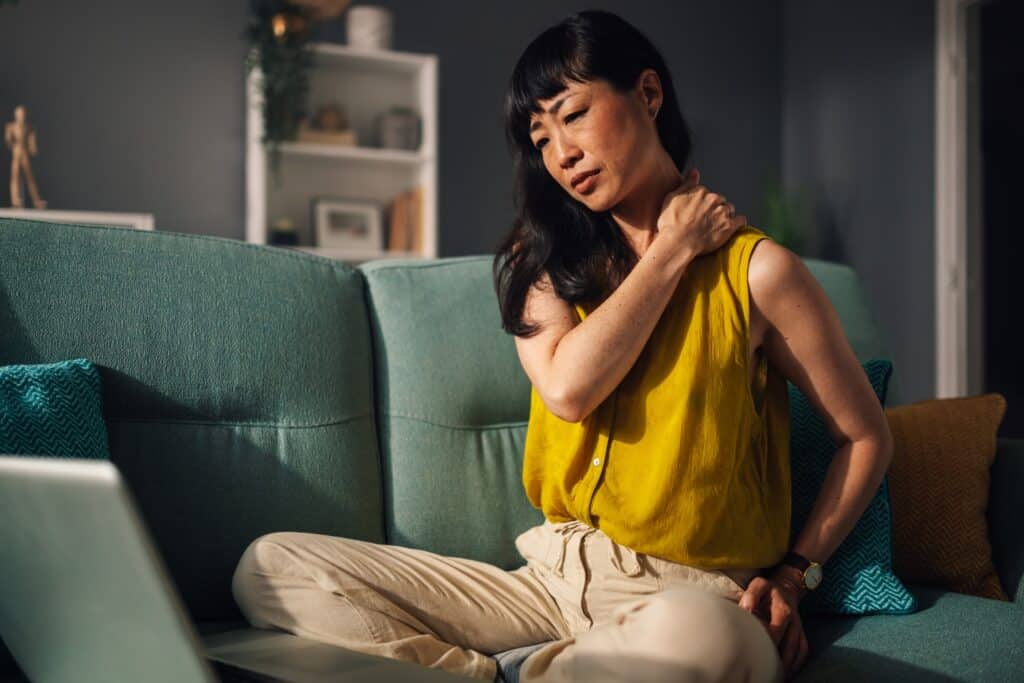Asian woman wearing a yellow top is seated cross-legged on a teal couch, holding her shoulder in apparent discomfort. She is next to a laptop and the home is warmly lit and decorated.